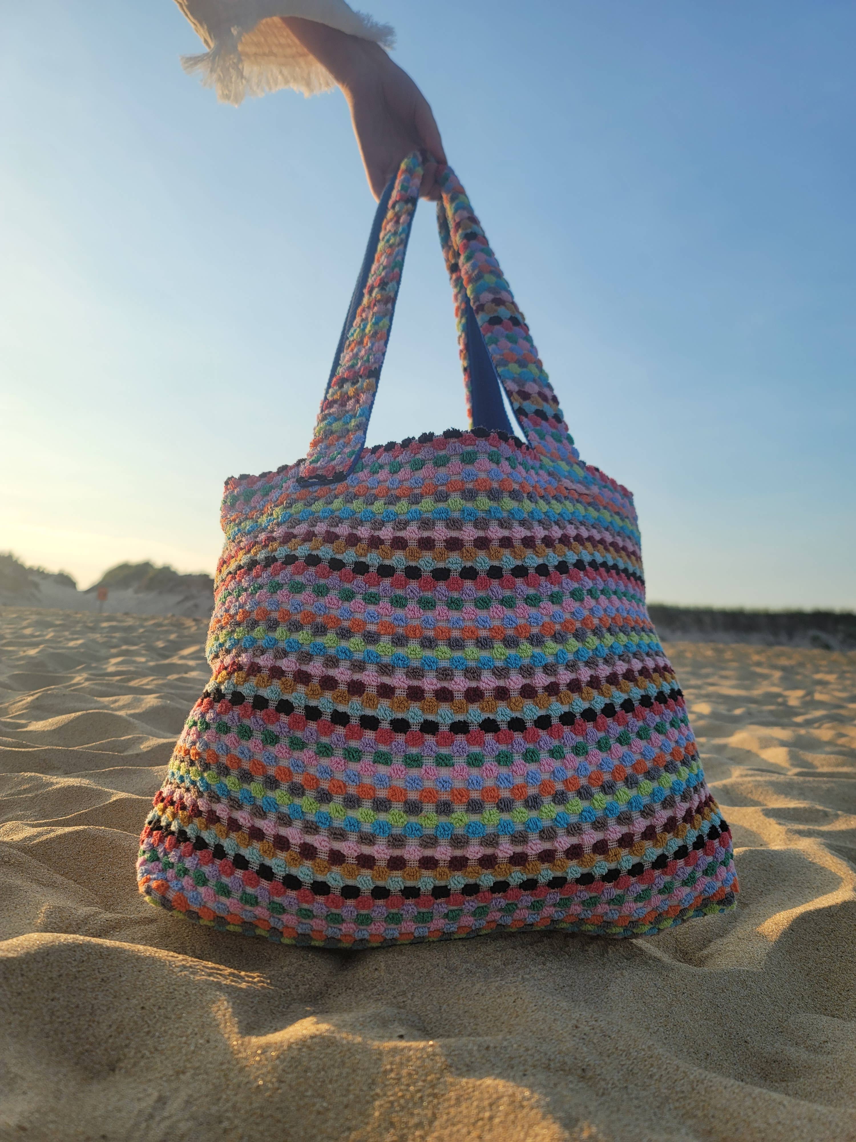 A multicolor handwoven tote bag with a geometric pattern, placed on a sandy surface under a clear sky.