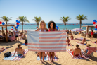 Two women holding a red and white striped towel on a beach with palm trees and ocean in the background.