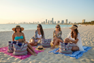 Four women sitting on a beach with colorful bags and a city skyline in the background